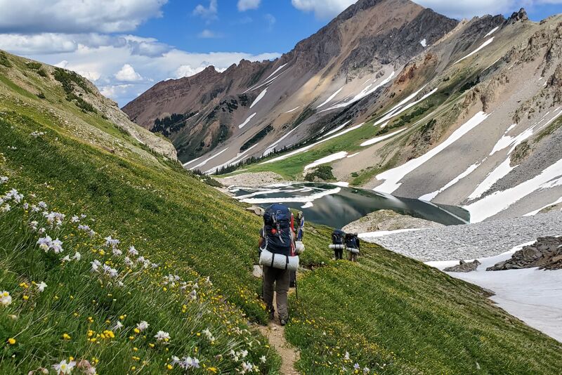 The image shows a scenic mountain landscape with three hikers on a trail. The hikers are walking away from the viewer, towards a lake nestled between the mountains. Patches of snow are visible on the mountain slopes, contrasting with the green grass and wildflowers in the foreground. The sky is partly cloudy, adding depth to the scene.